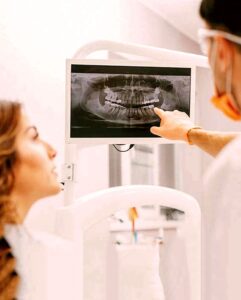 Clinical scene inside a dental surgery where a dentist wearing gloves points at a panoramic dental x-ray on a screen while a patient looks on, used at Smile Concepts in Sydney to explain bone levels, missing teeth, and implant options. The image highlights how we rely on detailed imaging before recommending any implant, graft, or extraction. It supports content about careful diagnosis, safe planning, and why thorough scans are vital for long-term implant stability and predictable treatment outcomes.